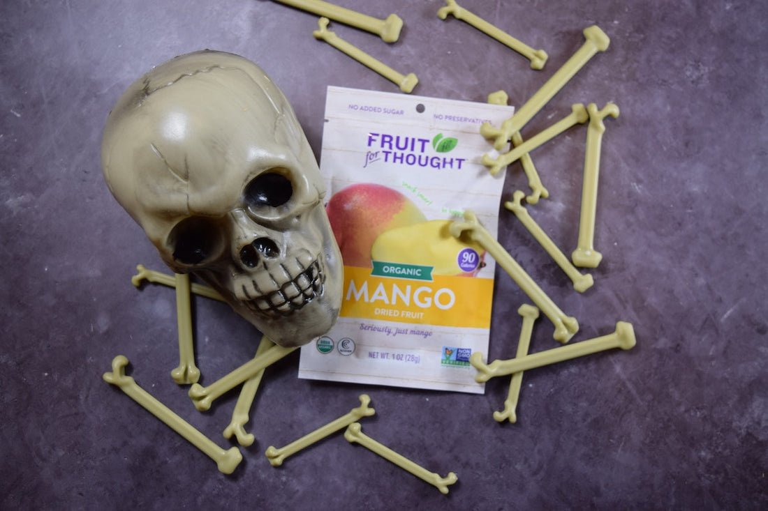 A fake skeleton head and bones lay next to a Fruit For Thought Organic Dried Mango snack pack.