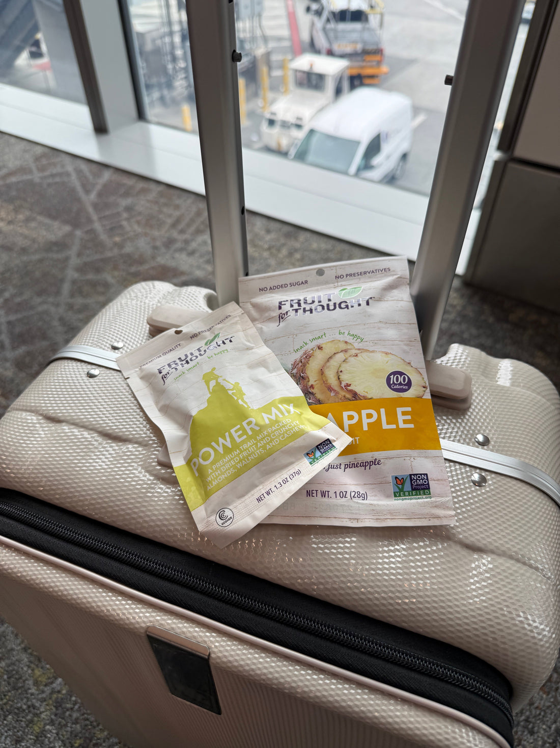 A white suitcase is positioned next to a window at an airport. On top of the suitcase are snack sized-bags of dried pineapple and power mix.
