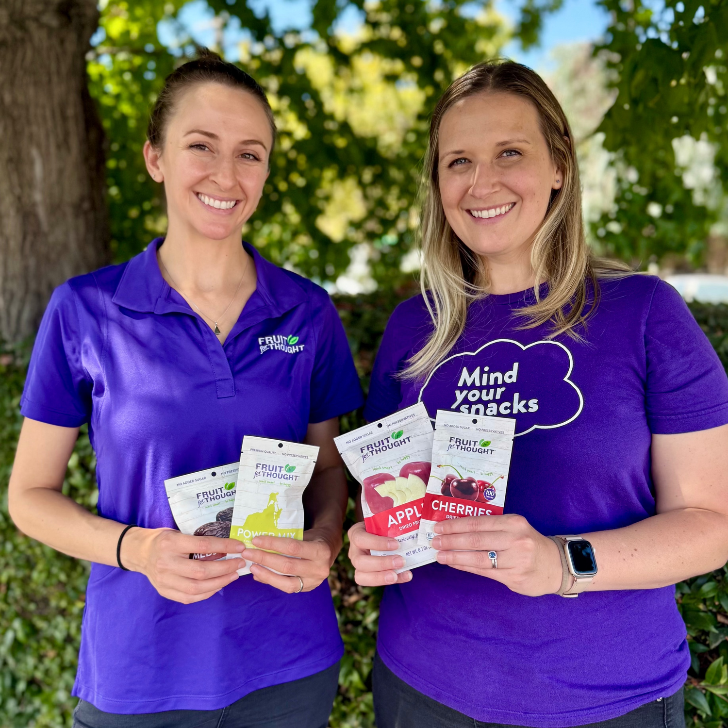 Two women in purple shirts holding fruit snacks outdoors.