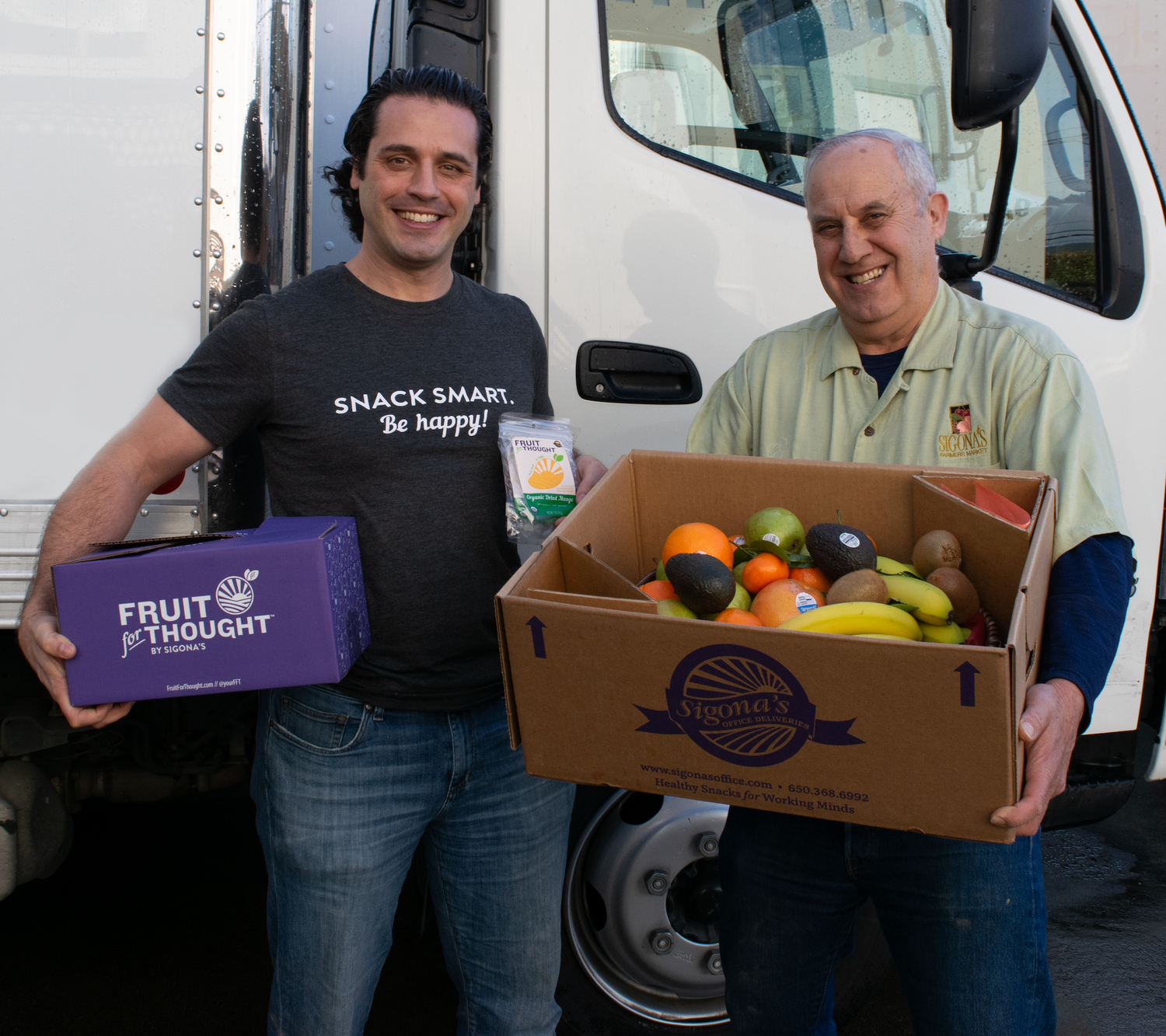 Two men holding a box of fruit and a box labeled 'Fruit of the Thought' in front of a white truck.