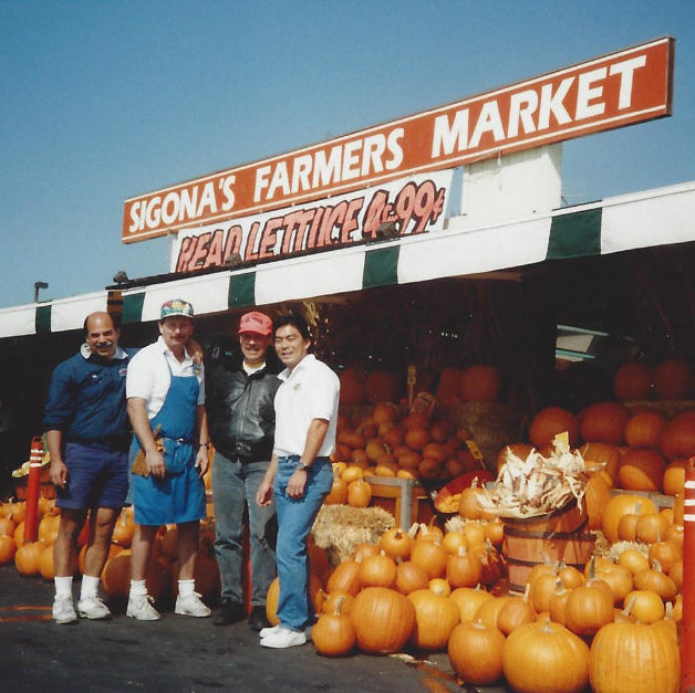 Three men standing in front of a farmers market with pumpkins and produce.